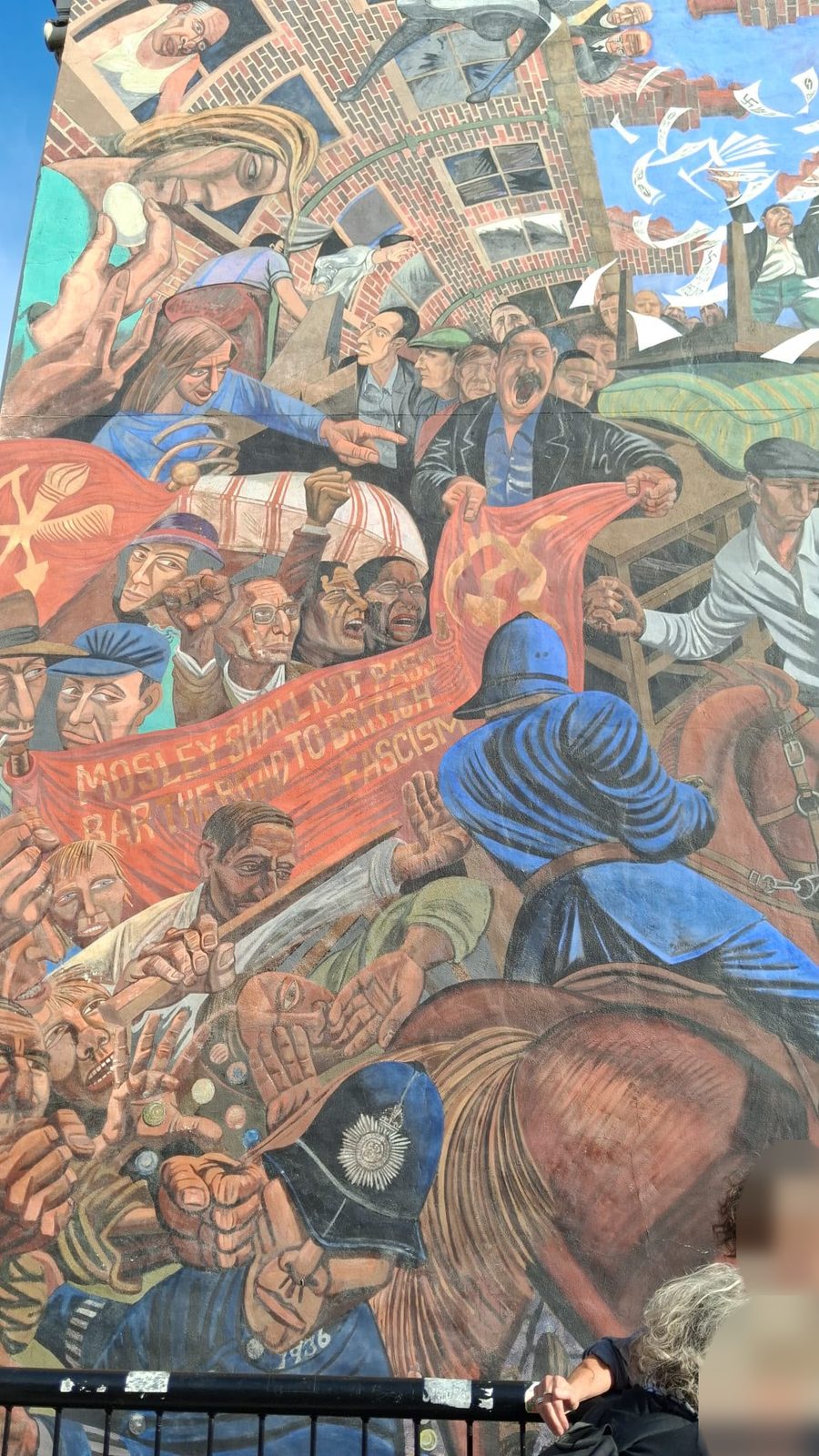 Detail of a painted mural: a crowd holds up a red banner reading “Mosley shall not pass. Bar the road to British fascism” as police officers push into them, one on horseback. Two people, faces blurred, stand in front of the mural.