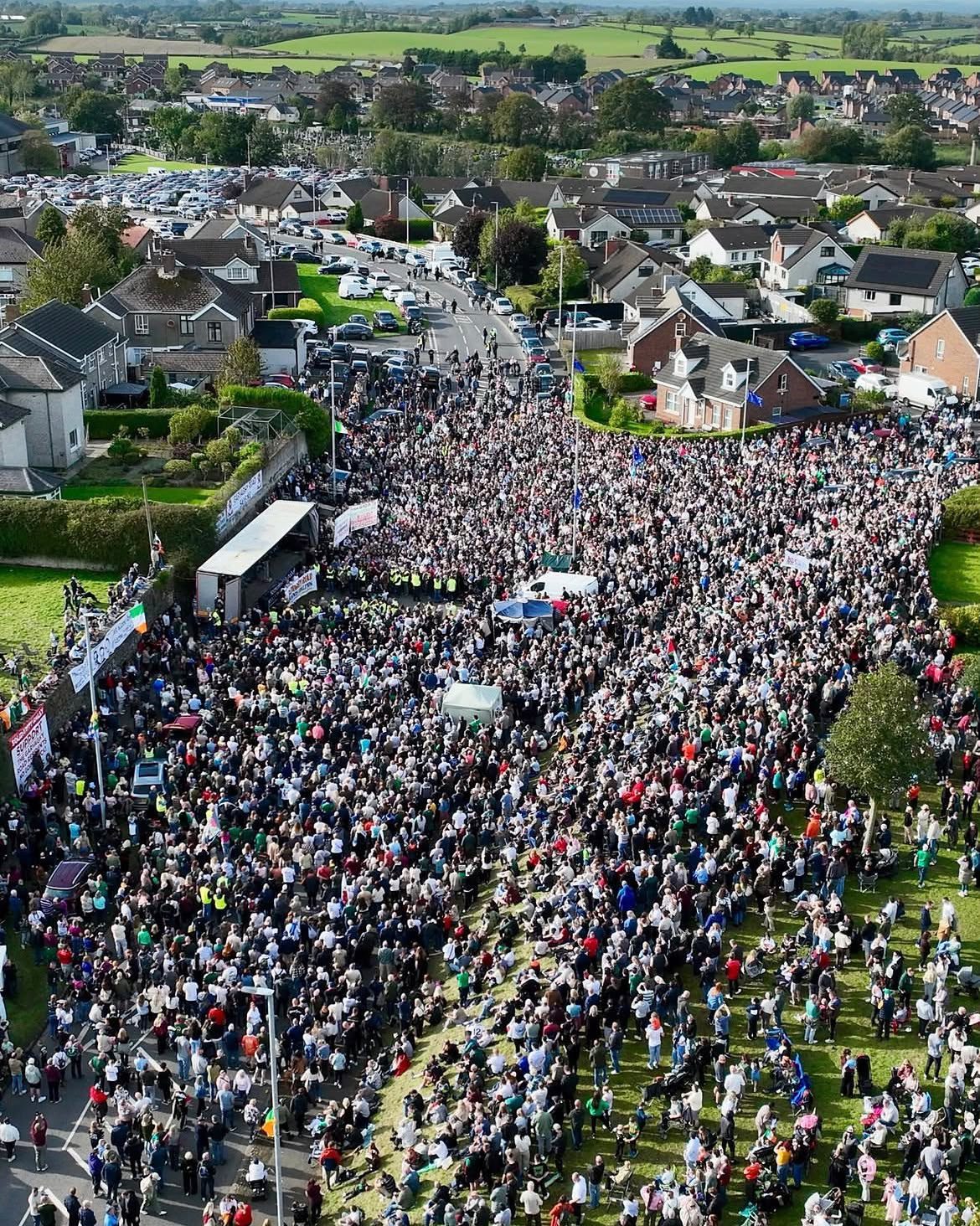Aerial view of a very large crowd filling a road and a grassy slope between houses, gathered around a stage on a lorry trailer, with flags and banners among them.