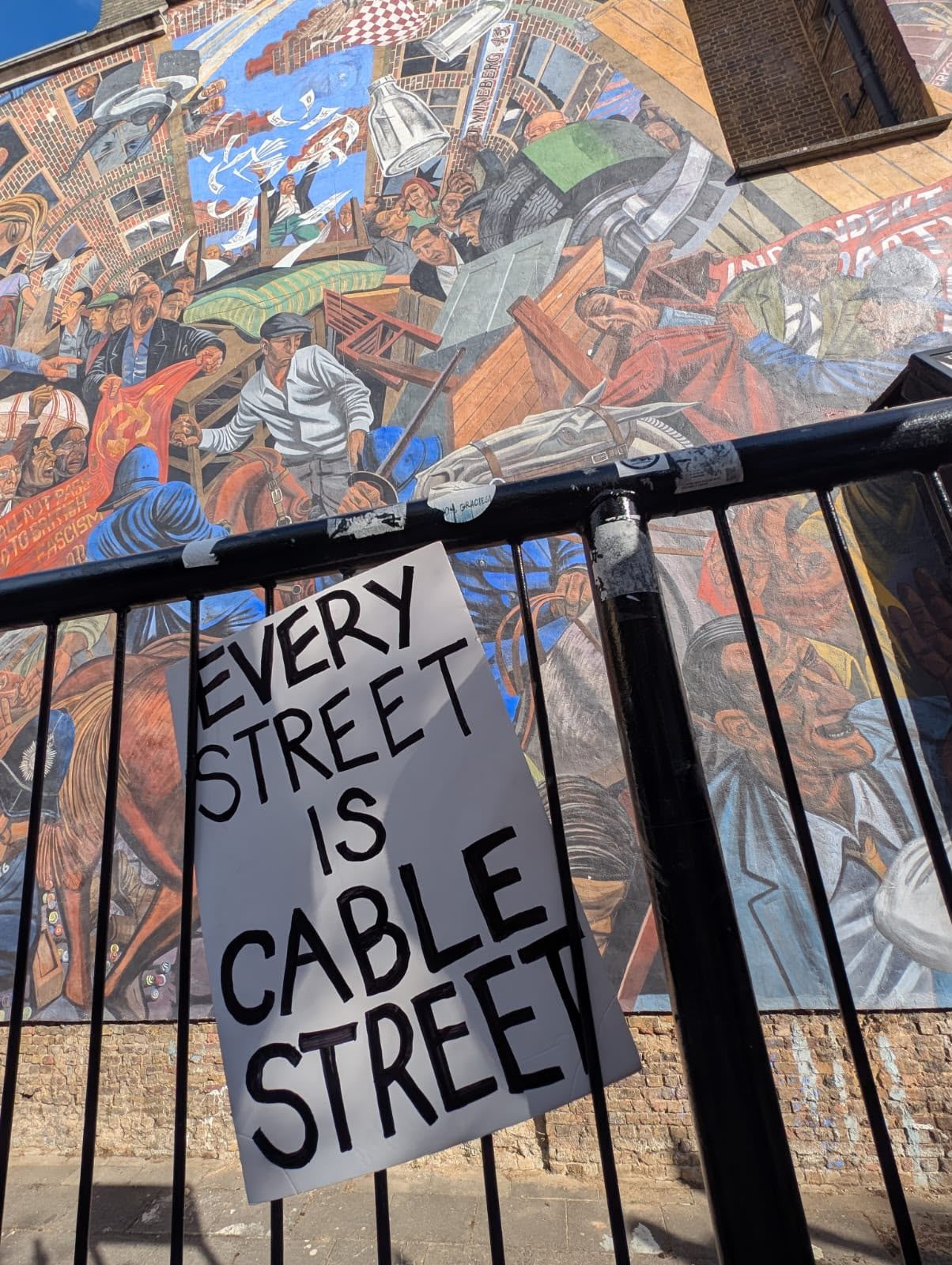 A hand-lettered placard reading “Every street is Cable Street” tied to black railings, in front of a large painted mural of a street battle with crowds, police and horses.